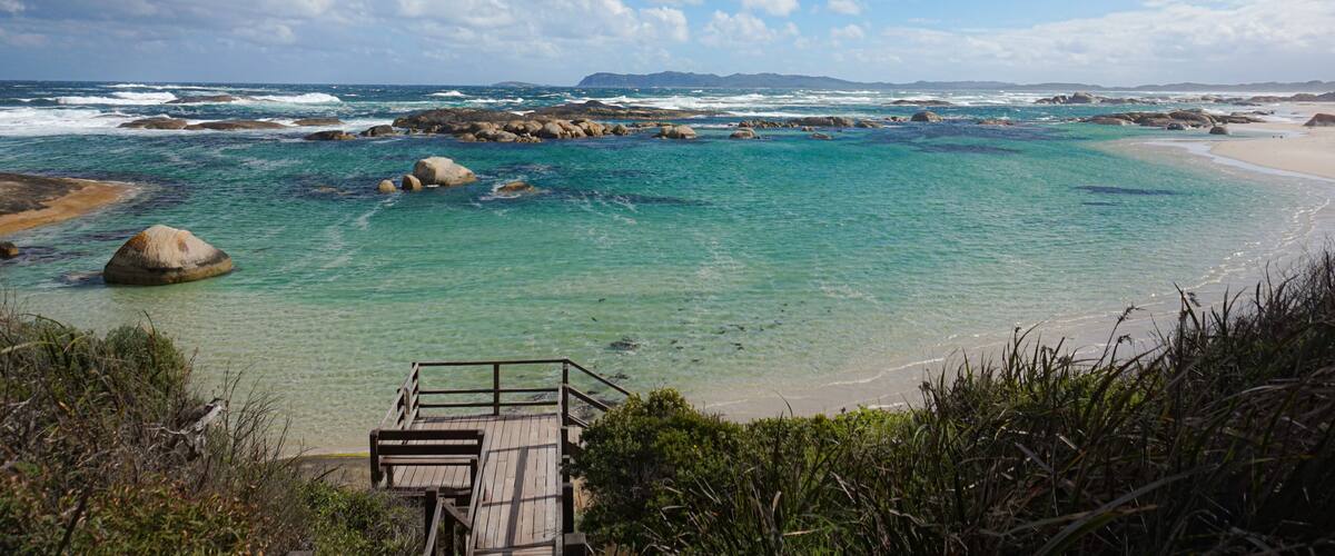 A coastal landscape with clear turquoise waters, rocky outcrops, and a wooden walkway leading to a sandy beach.