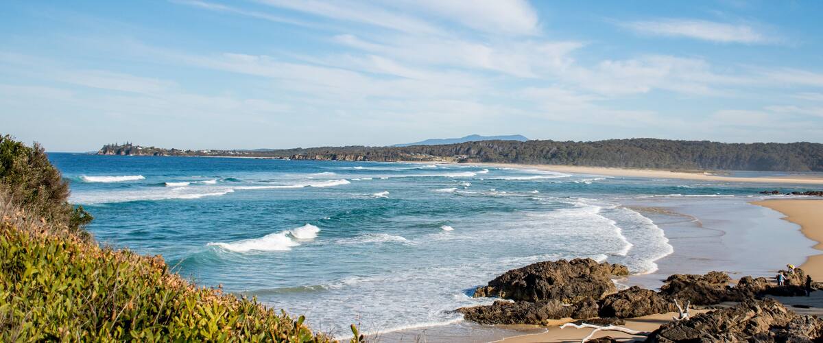 A coastal landscape with waves crashing onto a sandy beach, rocky outcrops, and a forested hill in the background.