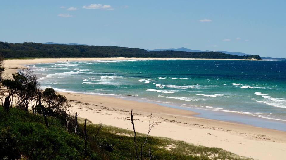A coastal landscape with a sandy beach, turquoise ocean, and a forested hill in the background.