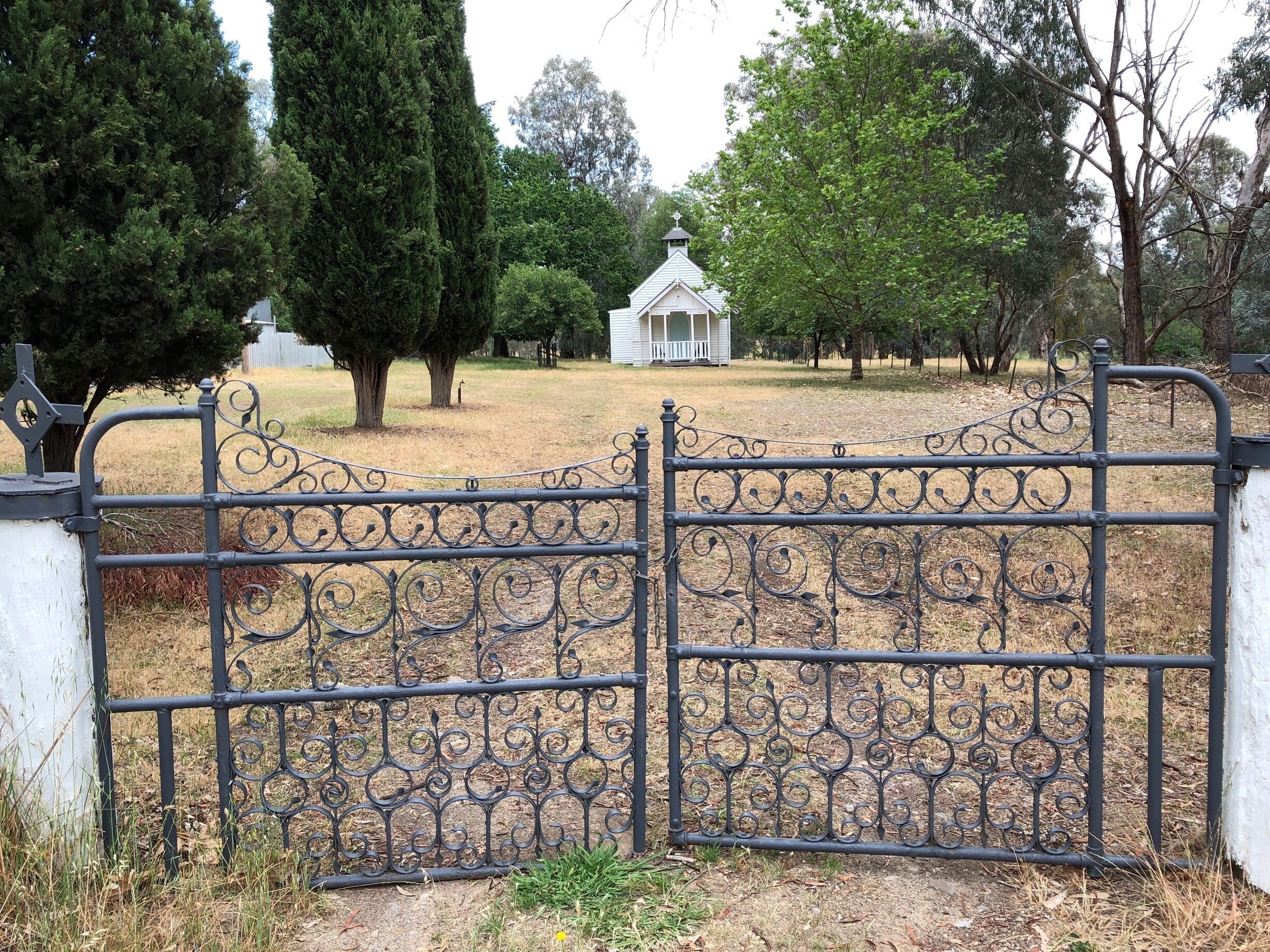 A wrought iron gate with intricate designs, a white building with a pointed roof in the background, and a grassy area with trees.
