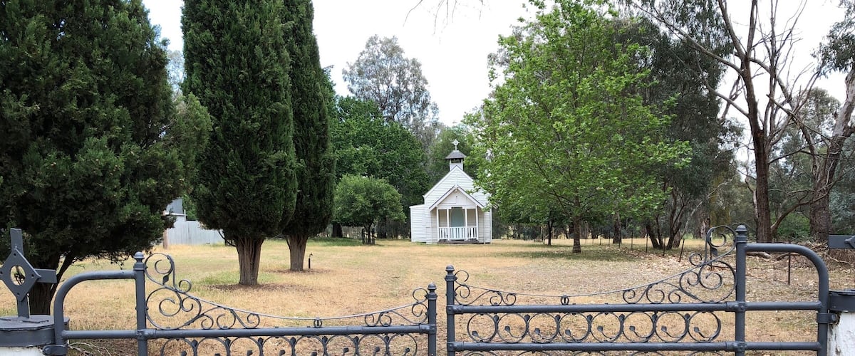 A wrought iron gate with intricate designs, a white building with a pointed roof in the background, and a grassy area with trees.