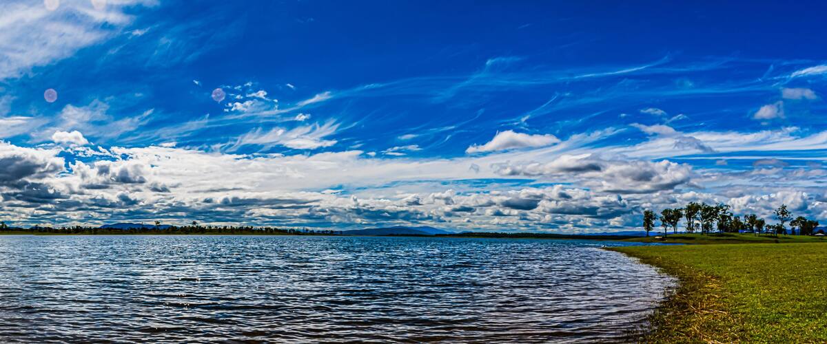 A lake with a grassy shoreline and a distant mountain range under a partly cloudy sky.