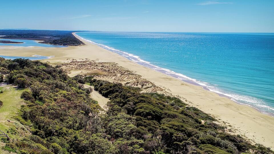 A coastal landscape with a sandy beach, turquoise waters, and dense green vegetation.