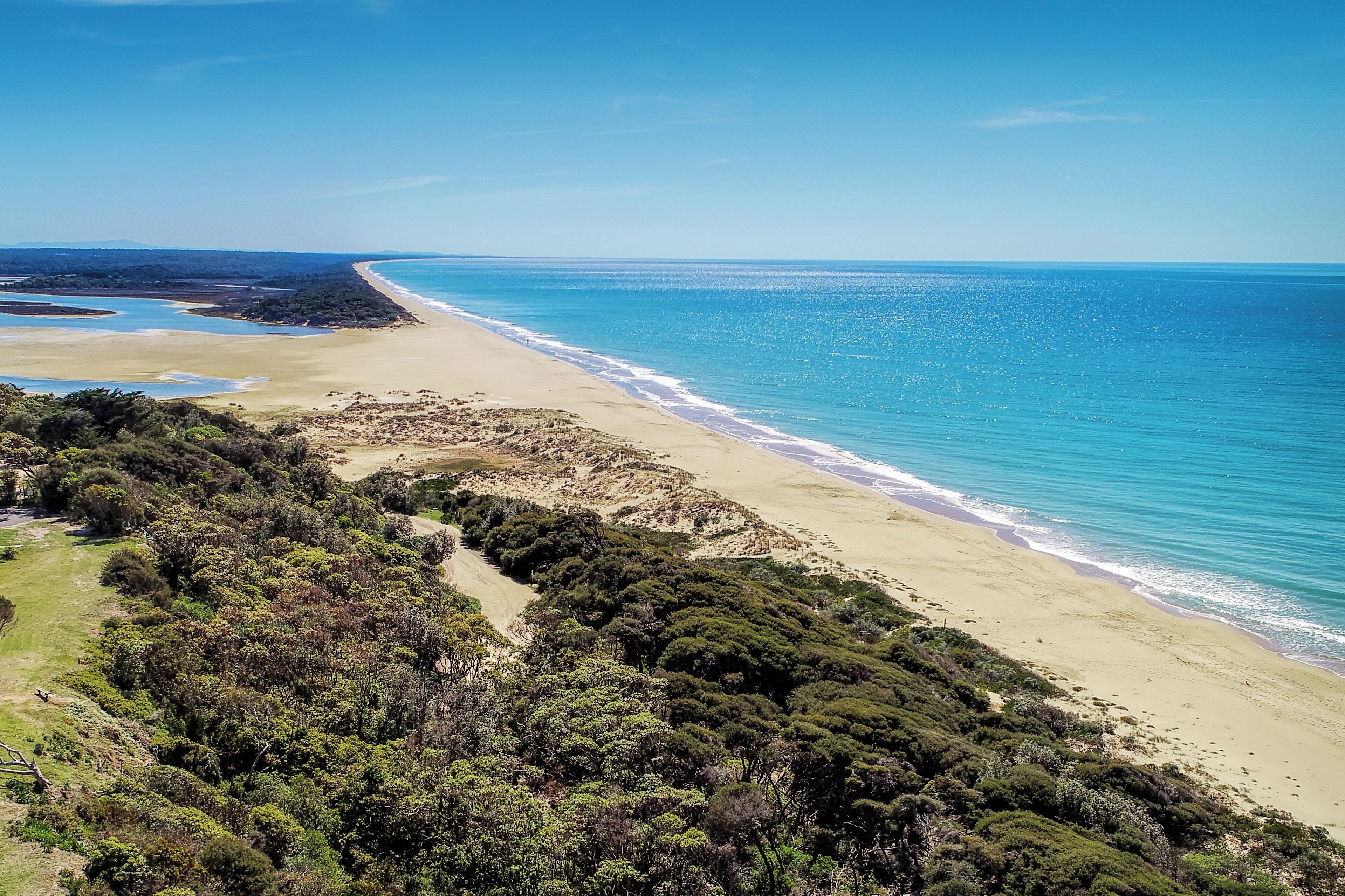 A coastal landscape with a sandy beach, turquoise waters, and dense green vegetation.