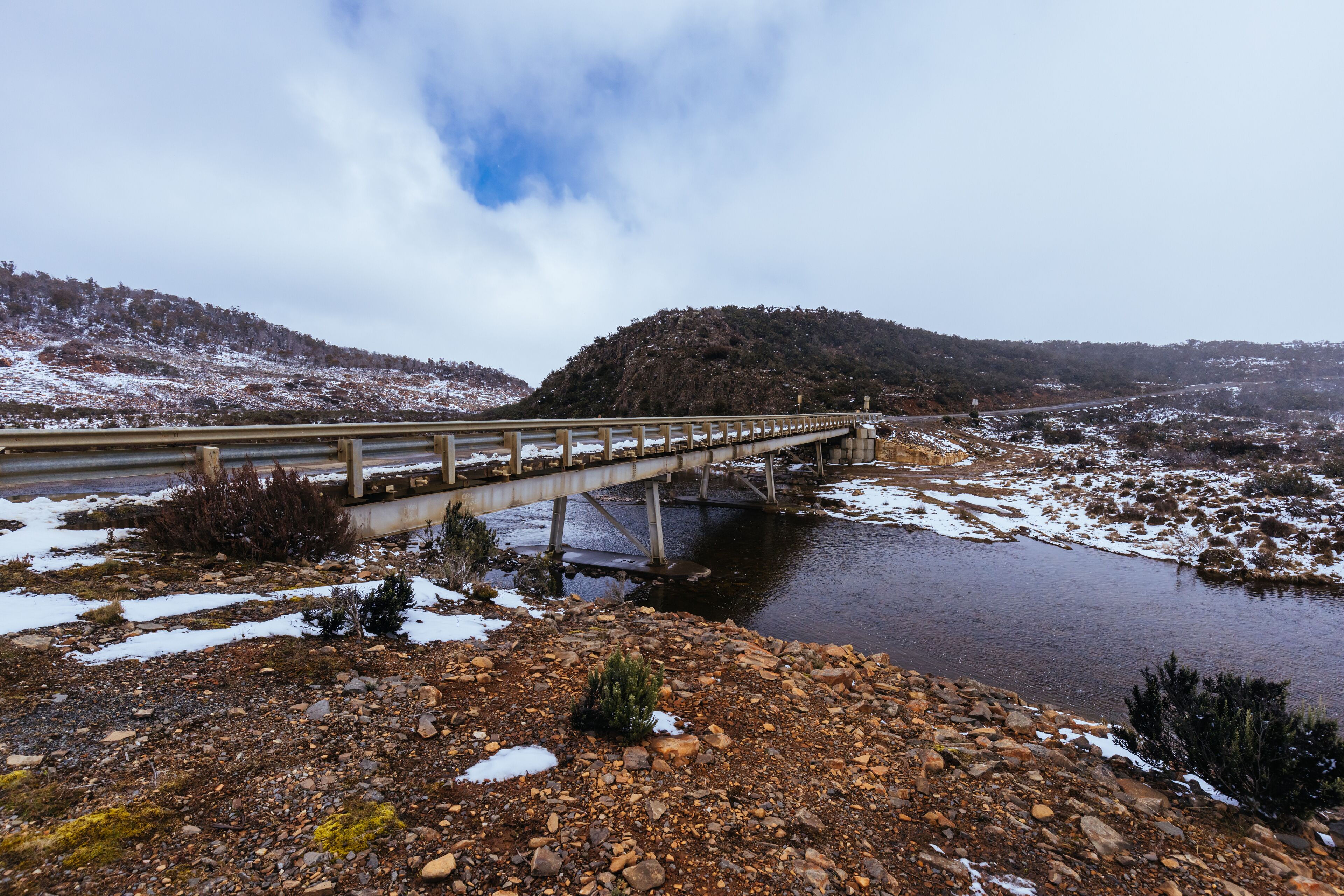 A bridge over a river with snow on the ground and hills in the background.