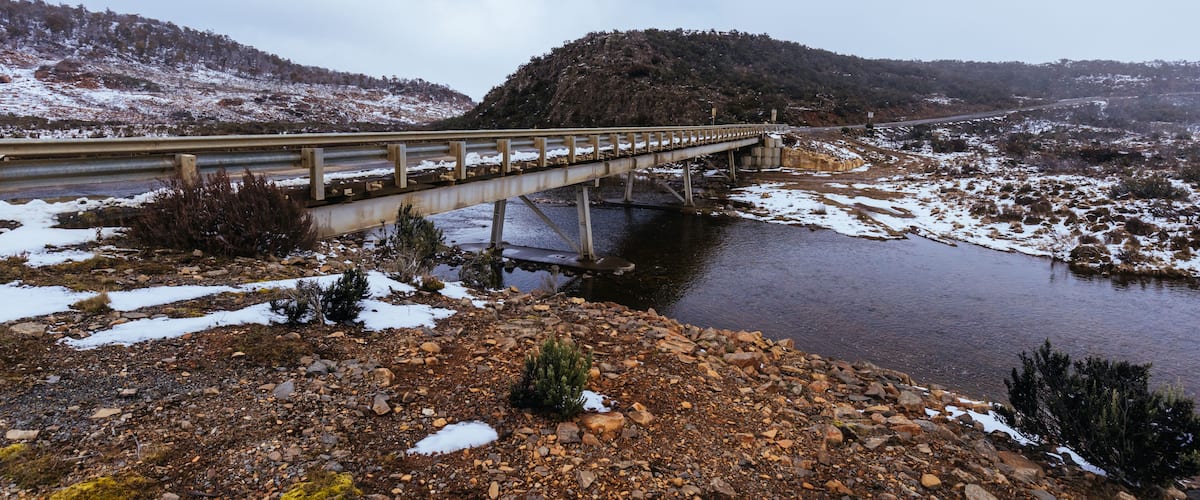 A bridge over a river with snow on the ground and hills in the background.