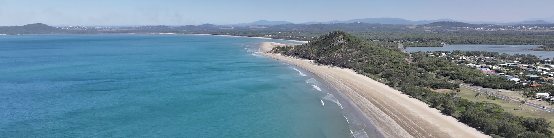 A coastal landscape with a sandy beach, clear blue waters, and a backdrop of hills and a small town.