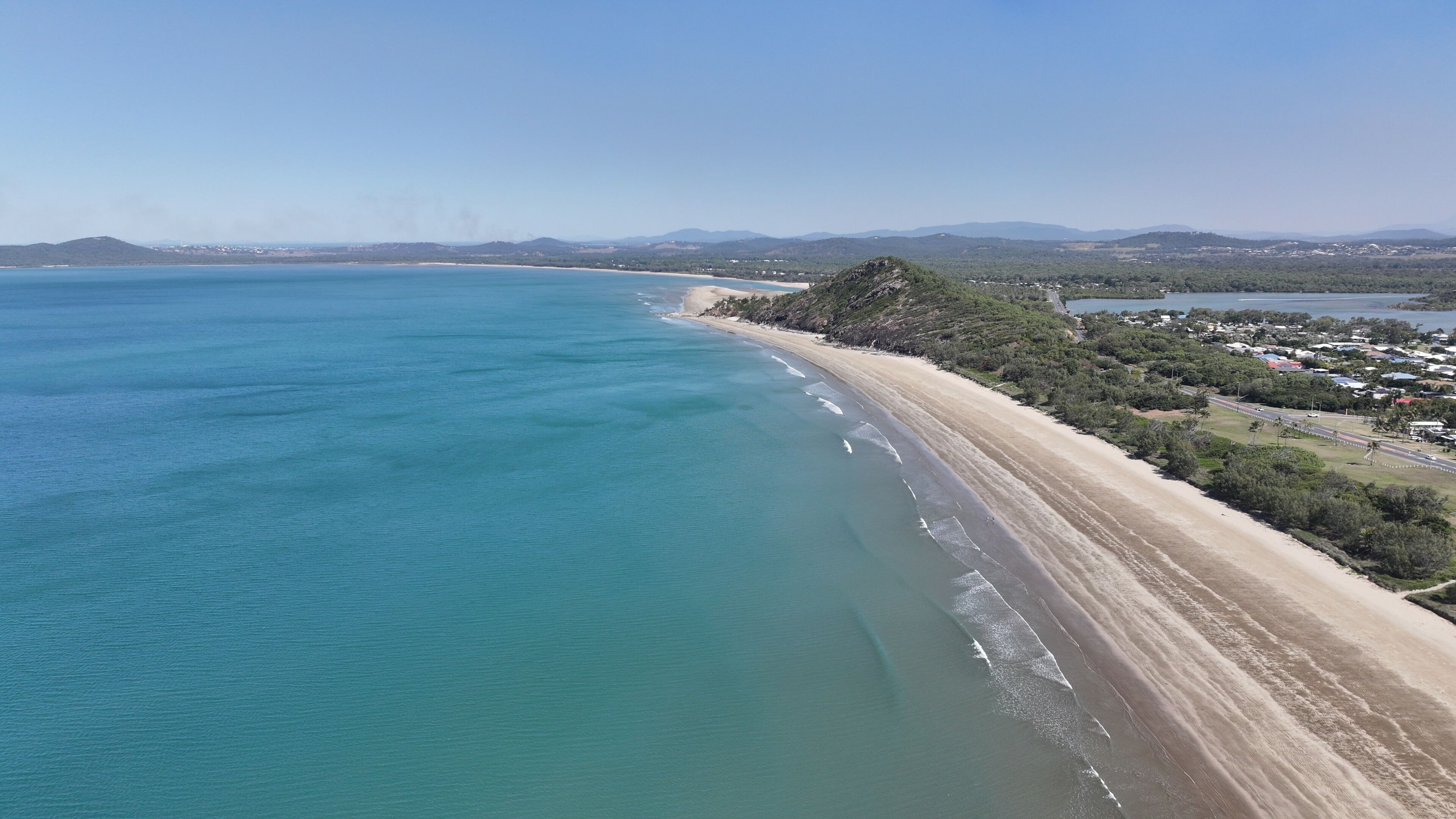 A coastal landscape with a sandy beach, clear blue waters, and a backdrop of hills and a small town.