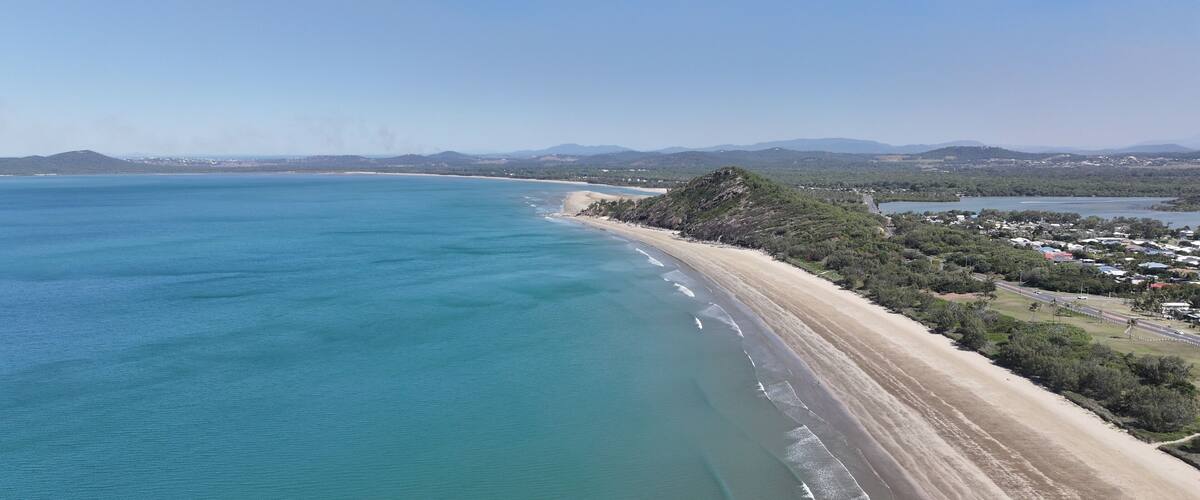 A coastal landscape with a sandy beach, clear blue waters, and a backdrop of hills and a small town.