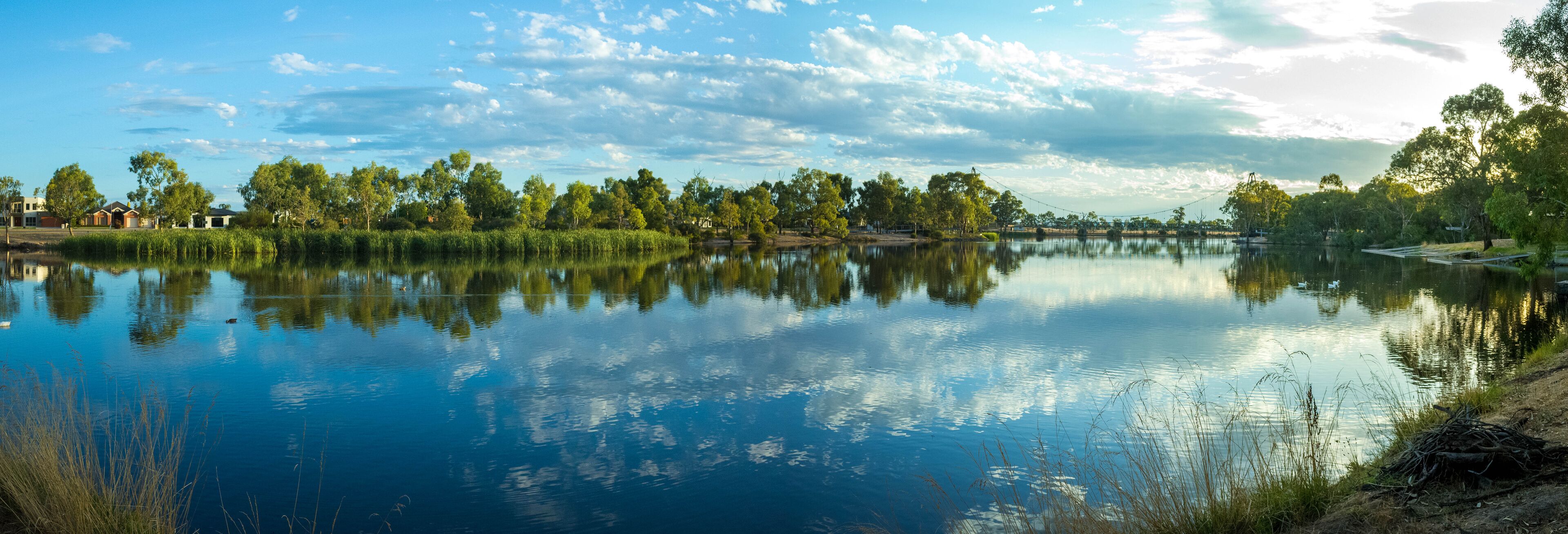Panoramic view of Wimmera River. Riverbank with water reflection of beautiful cloudy sky, residential houses and Anzac Centenary Bridge in the distance. Horsham, VIC Australia.