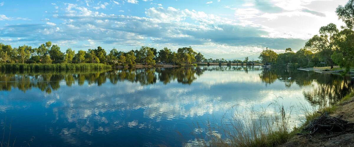 Panoramic view of Wimmera River. Riverbank with water reflection of beautiful cloudy sky, residential houses and Anzac Centenary Bridge in the distance. Horsham, VIC Australia.