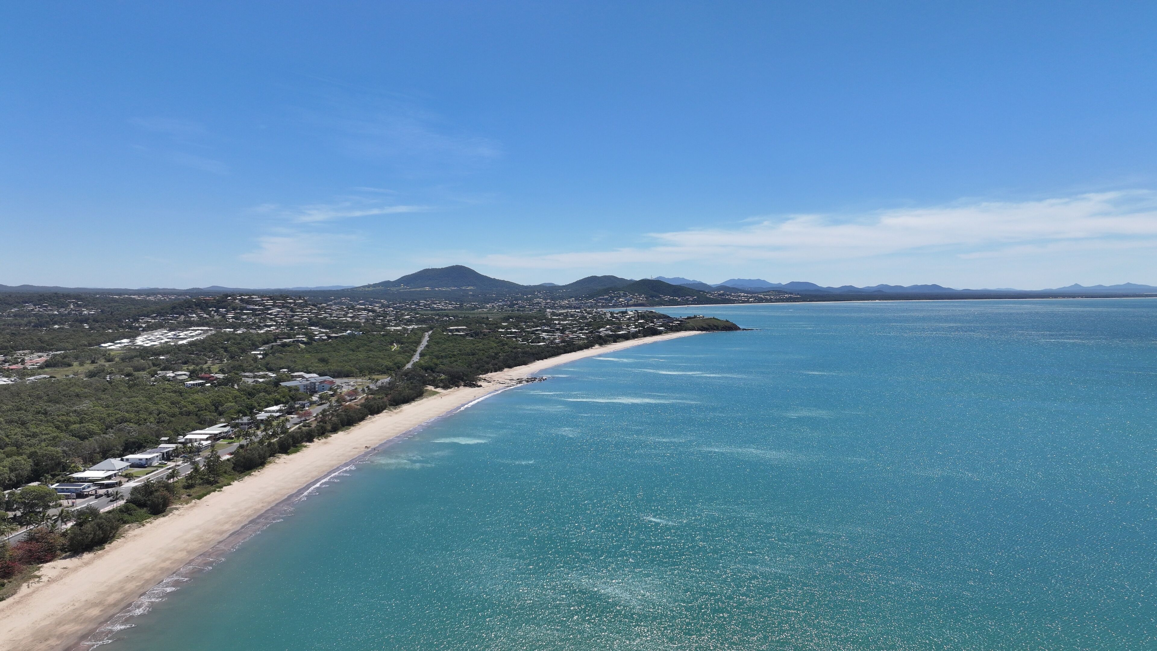 A coastal town with a beach, clear blue sea, and distant mountains.