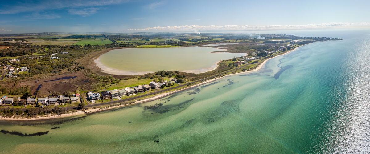 An aerial view of a coastal area with a large lake, a beach, and a residential area with houses and greenery.