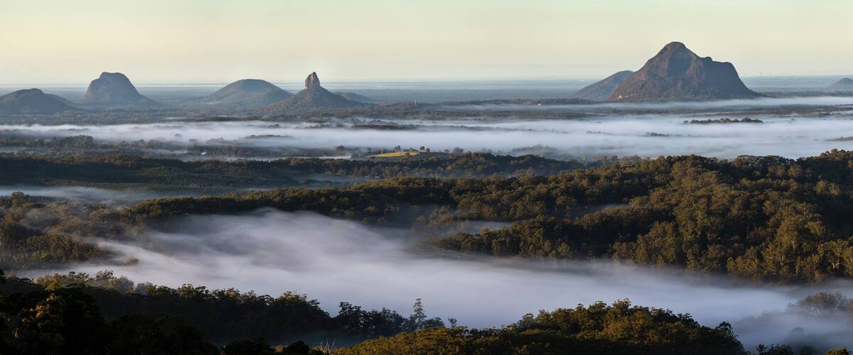 A landscape with a forested valley, mist-covered mountains, and a distant view of a coastline.