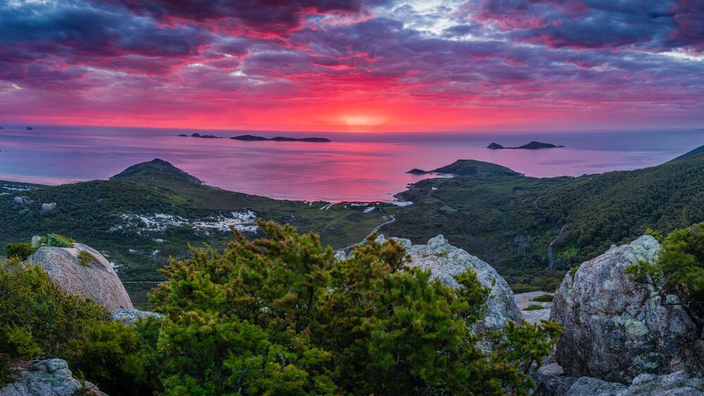 Eine Berglandschaft mit Sonnenuntergang über einem Gewässer, umgeben von dichten Wäldern und felsigen Felsen.