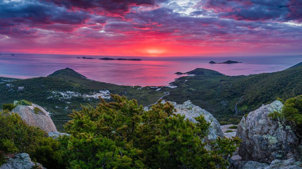 A mountainous landscape with a sunset over a body of water, surrounded by dense forests and rocky outcrops.