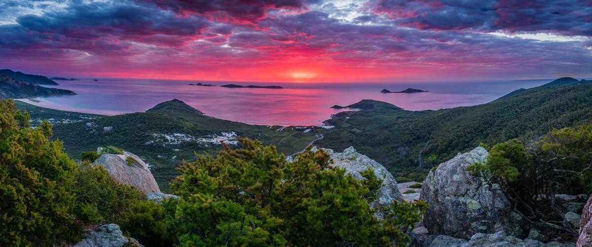 A mountainous landscape with a sunset over a body of water, surrounded by dense forests and rocky outcrops.