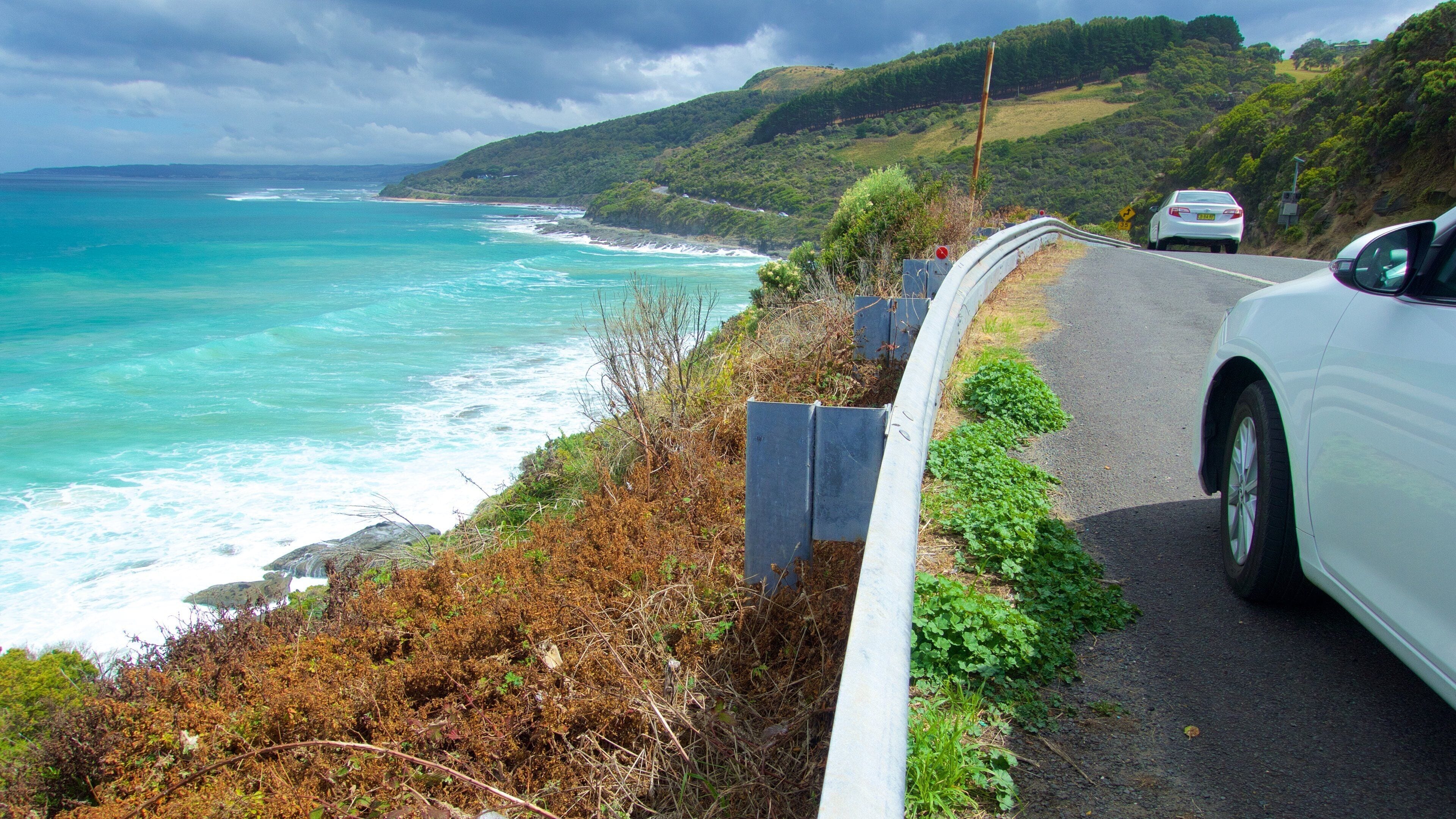 A coastal road with a white car driving along it, overlooking the ocean and green hills.