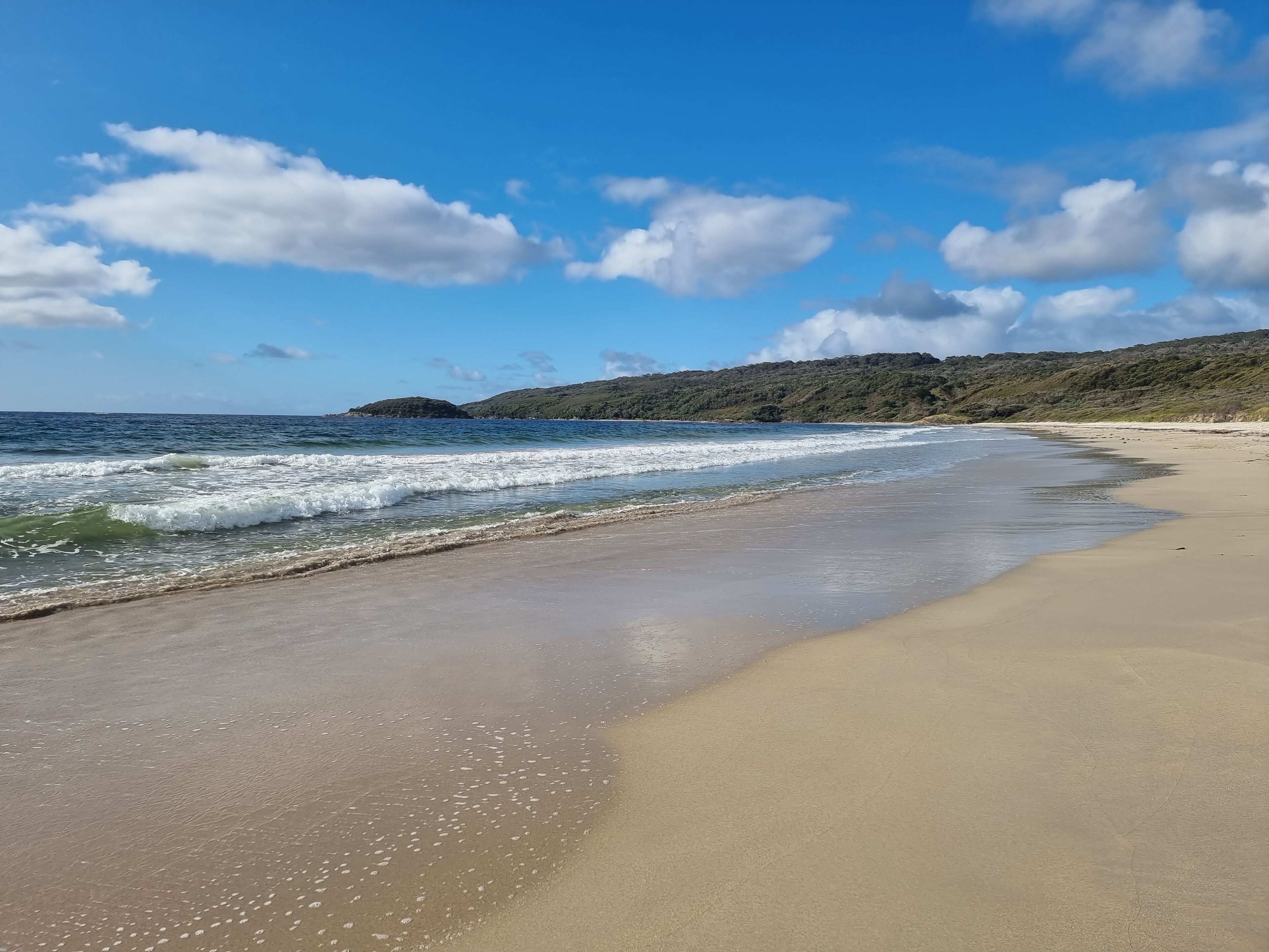 A sandy beach with gentle waves, a clear blue sky, and distant hills.