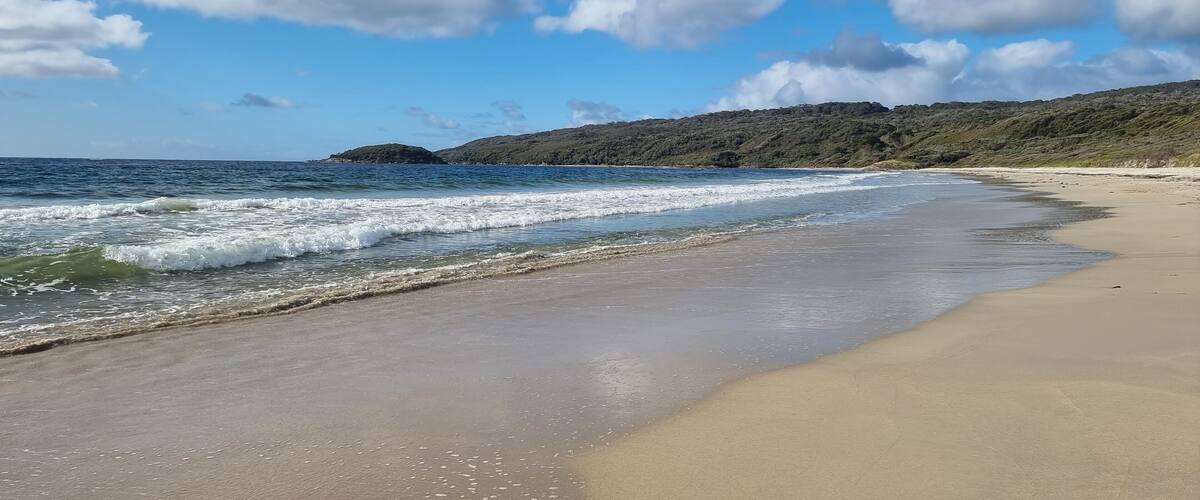 A sandy beach with gentle waves, a clear blue sky, and distant hills.