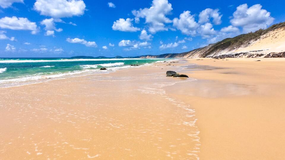A sandy beach with gentle waves, a clear blue sky, and distant mountains.