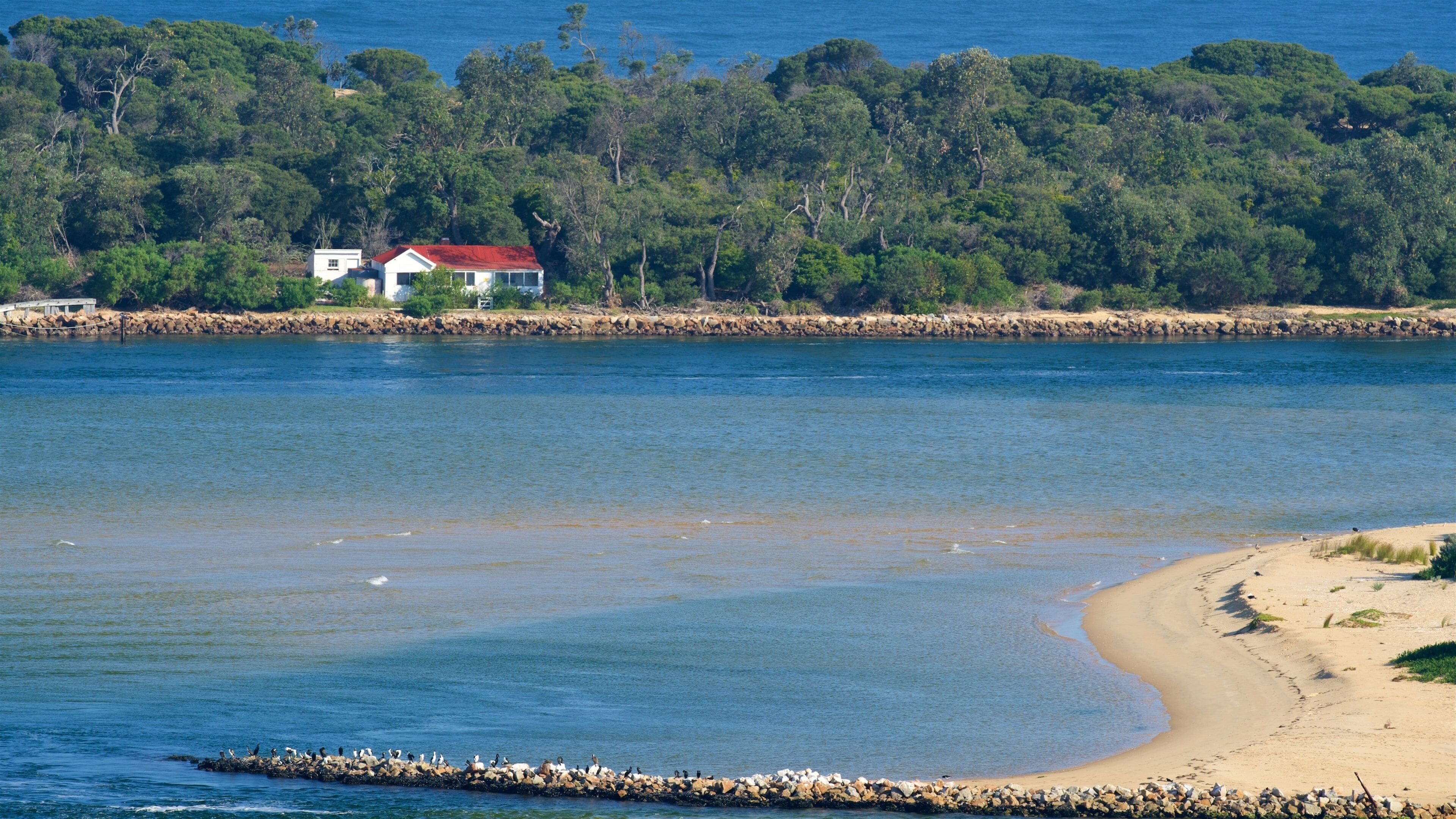 A coastal landscape with a sandy beach, calm water, a rocky breakwater, and a house with a red roof.
