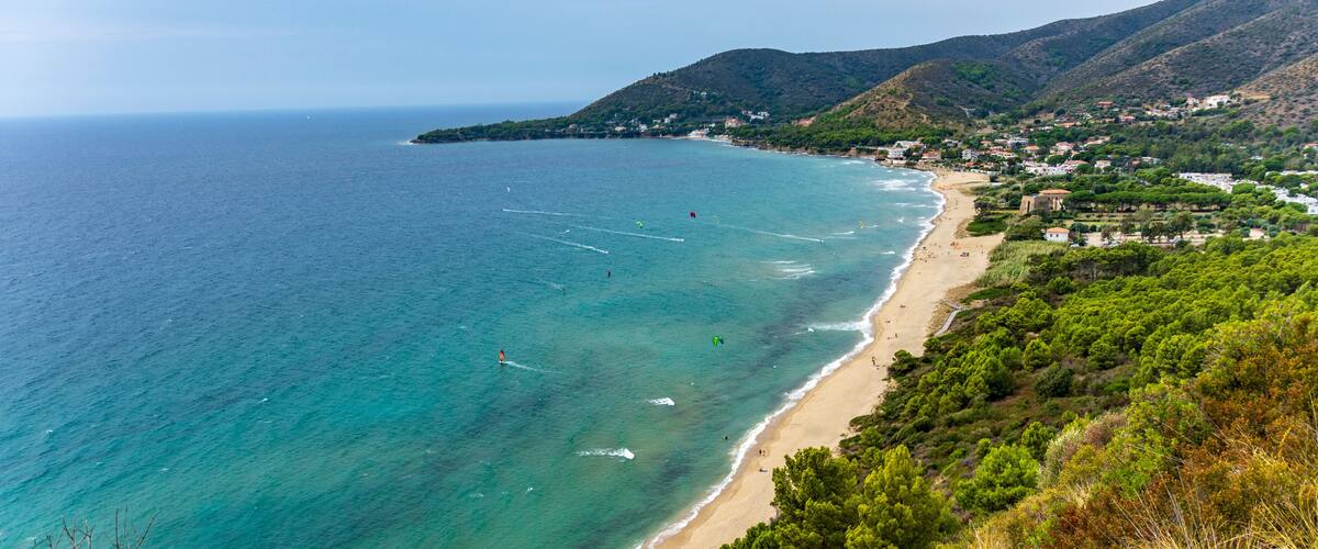 A coastal landscape with a beach, clear blue sea, and green hills.