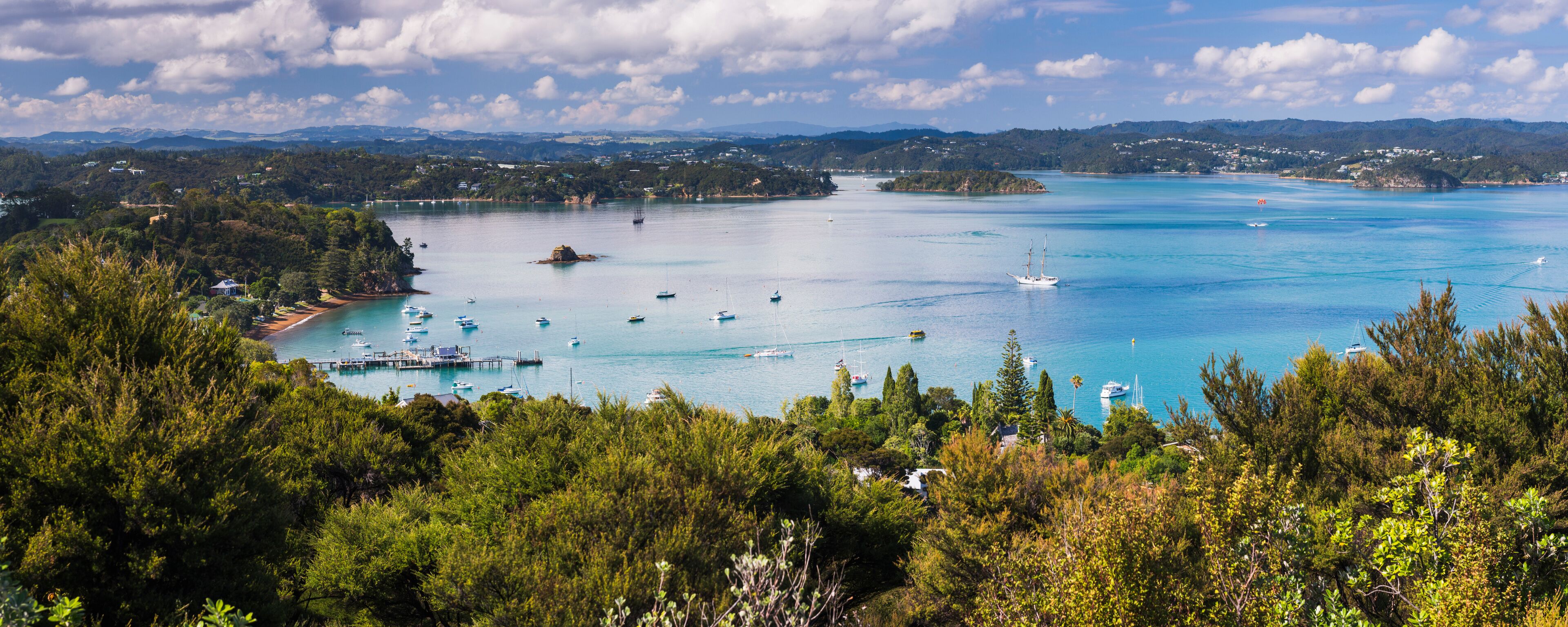 A coastal bay with clear turquoise waters, numerous boats, and lush green hills in the background.