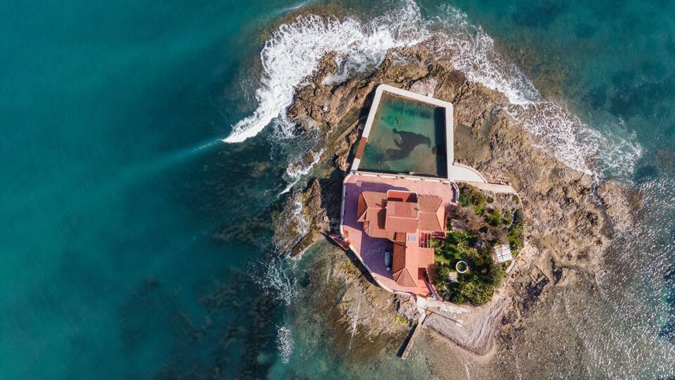 An aerial view of a coastal property with a swimming pool, a red-roofed building, and rocky terrain.