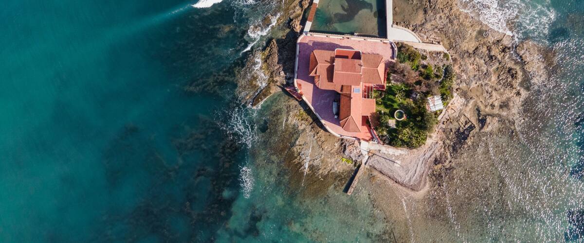 An aerial view of a coastal property with a swimming pool, a red-roofed building, and rocky terrain.