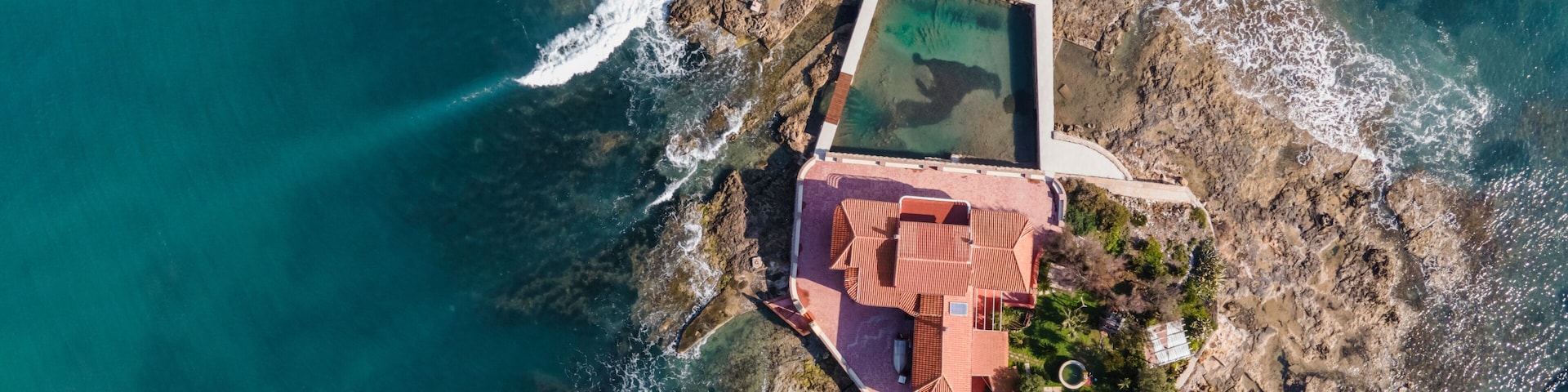 An aerial view of a coastal property with a swimming pool, a red-roofed building, and rocky terrain.