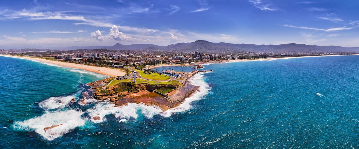 An aerial view of a coastal city with a prominent beach, rocky outcrops, and distant mountains.