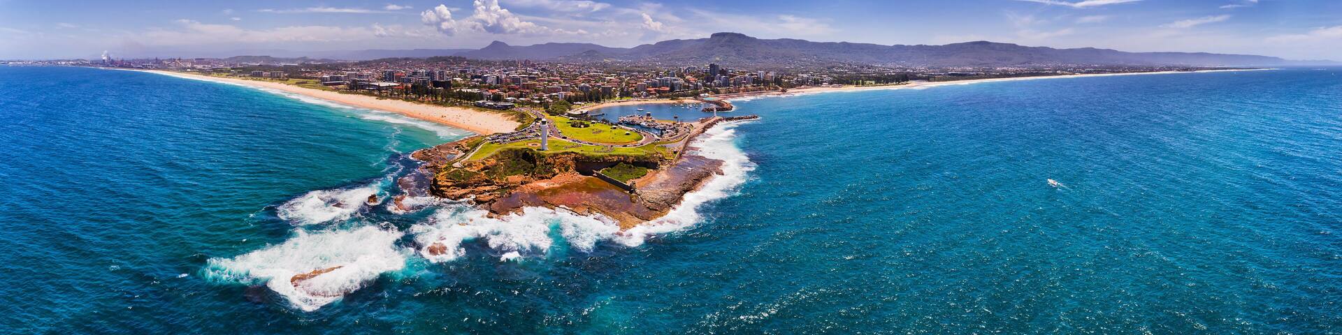 An aerial view of a coastal city with a prominent beach, rocky outcrops, and distant mountains.