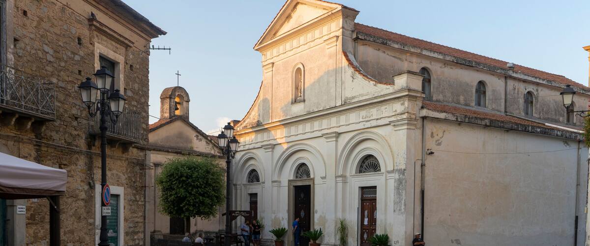 A historic church with a bell tower and arched windows, surrounded by cobblestone streets and potted plants.