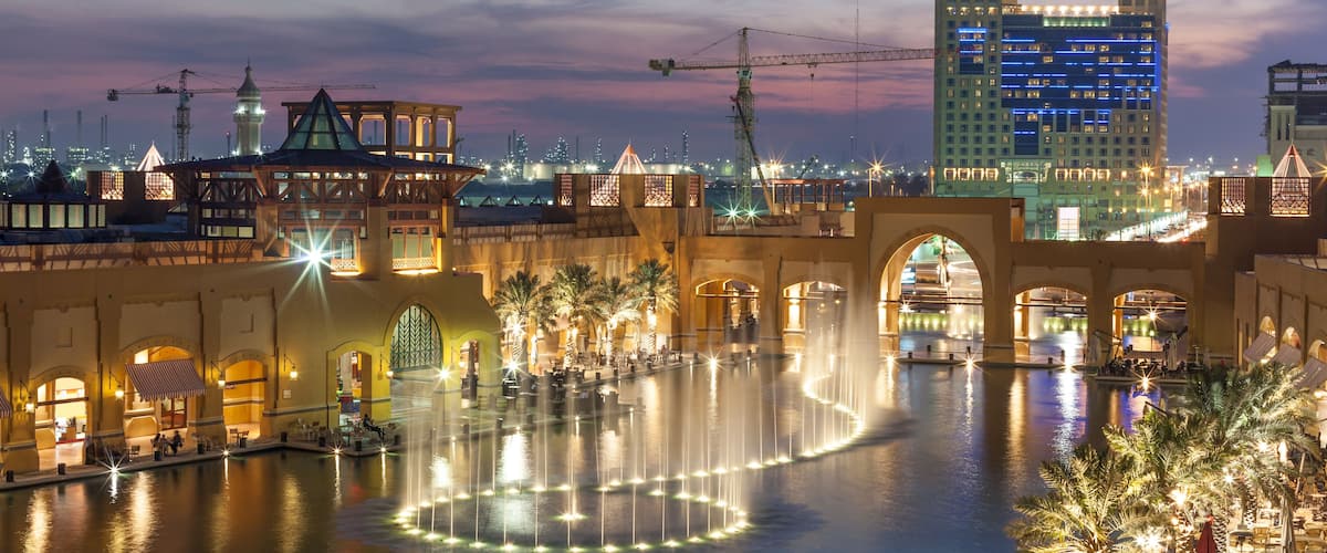 A nighttime cityscape with a water fountain, illuminated buildings, and a clear sky.
