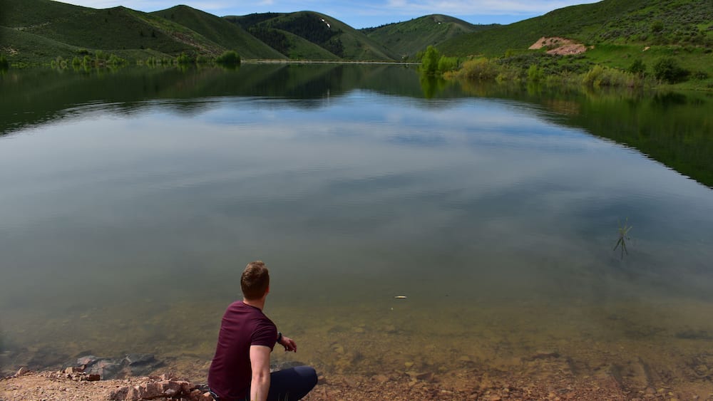 A calm lake with a person sitting on the shore, surrounded by green hills.