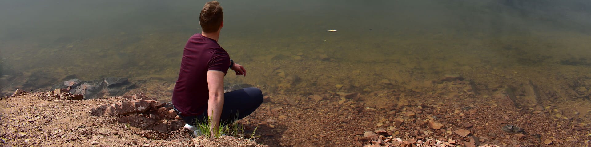 A calm lake with a person sitting on the shore, surrounded by green hills.