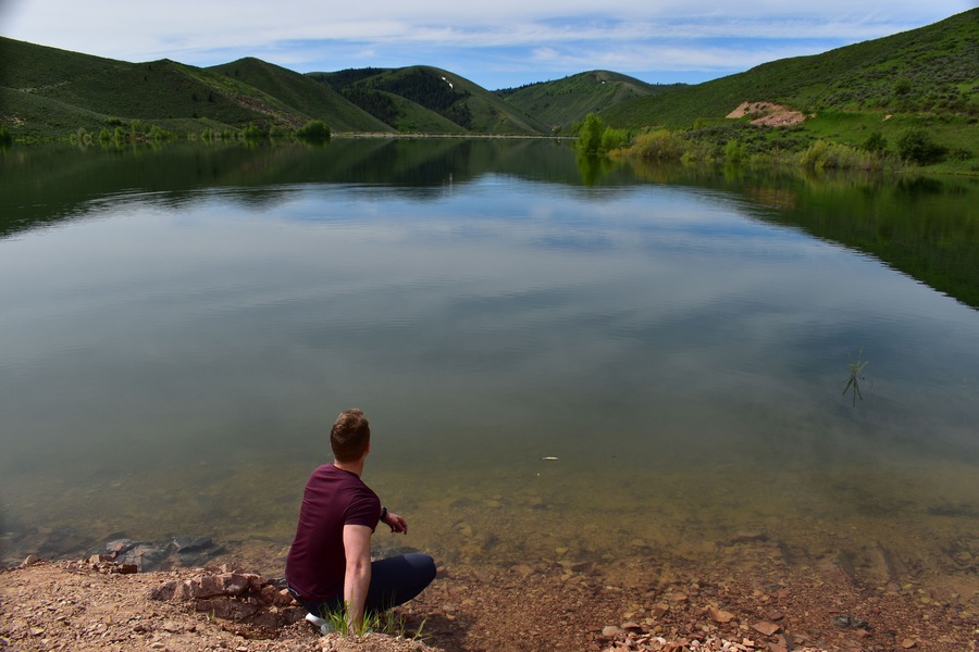 Um lago tranquilo com uma pessoa sentada na margem, rodeada por colinas verdes.
