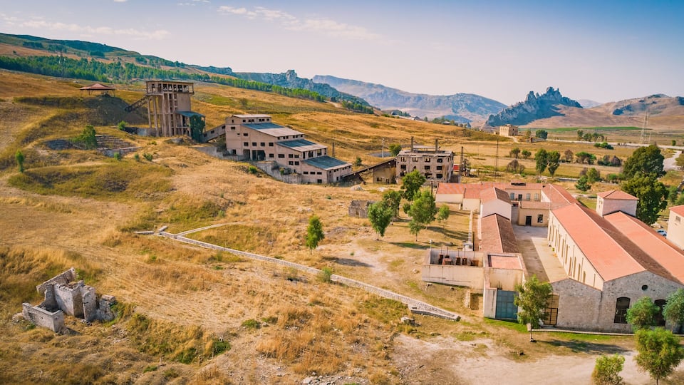 An abandoned industrial complex with buildings and structures amidst a hilly landscape.