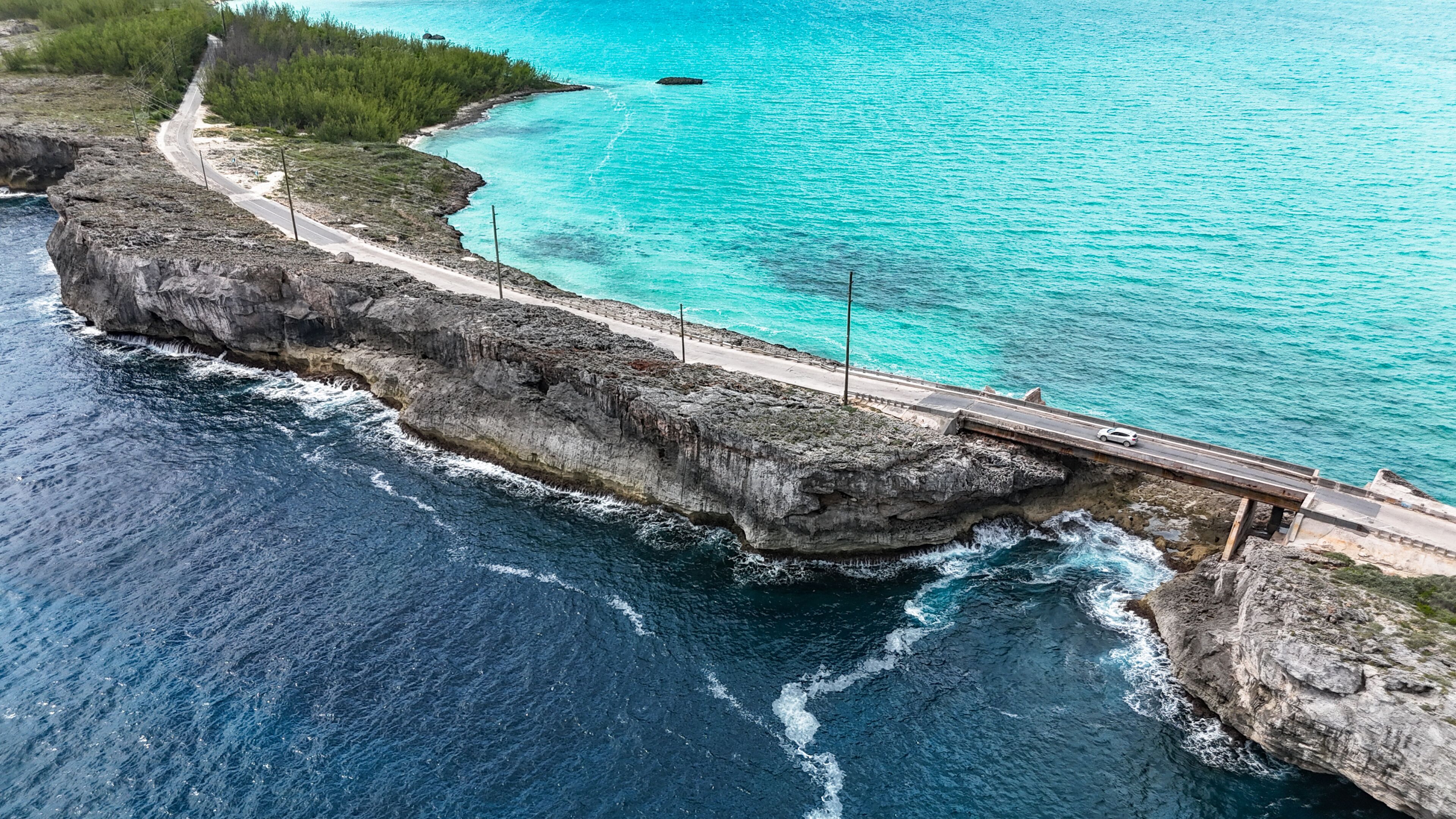 Aerial view of the Glass Window Bridge with a white car spanning a narrow rocky strip between the dark Atlantic Ocean and turquoise Caribbean waters in North Eleuthera, North Eleuthera, The Bahamas.