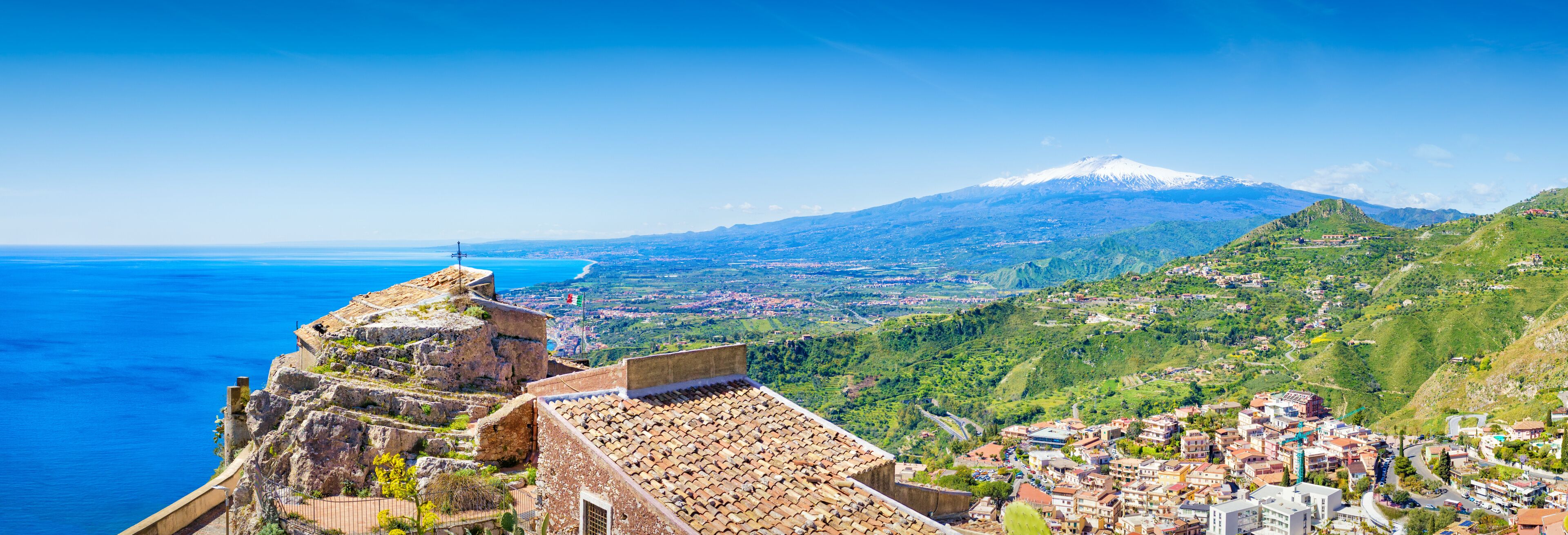 A coastal town with a historic fort overlooking the sea, a snow-capped mountain in the background, and lush green hills.