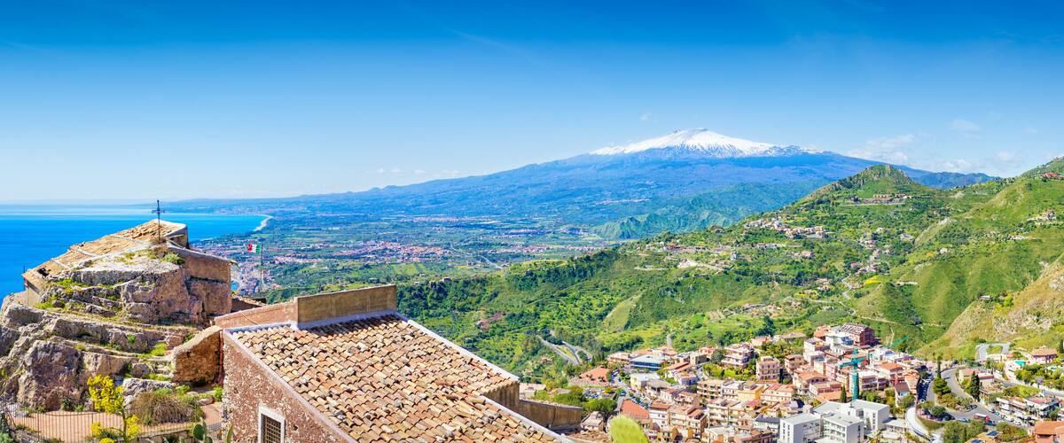 A coastal town with a historic fort overlooking the sea, a snow-capped mountain in the background, and lush green hills.