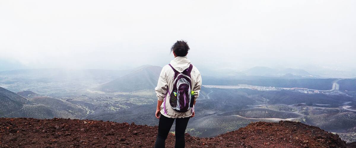 A person standing on a volcanic landscape with a view of a valley and distant mountains.