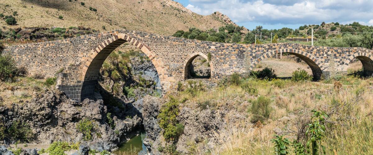 An old stone bridge with three arches over a rocky riverbed.