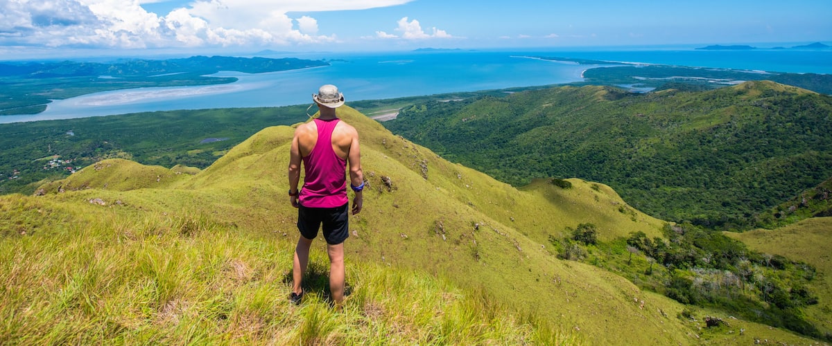 A person standing on a grassy hill overlooking a coastal landscape with a body of water and distant islands.