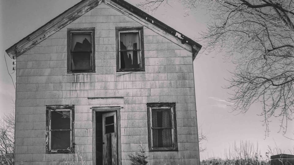 A weathered wooden house with a gabled roof and a chimney, surrounded by overgrown grass and a leafless tree.