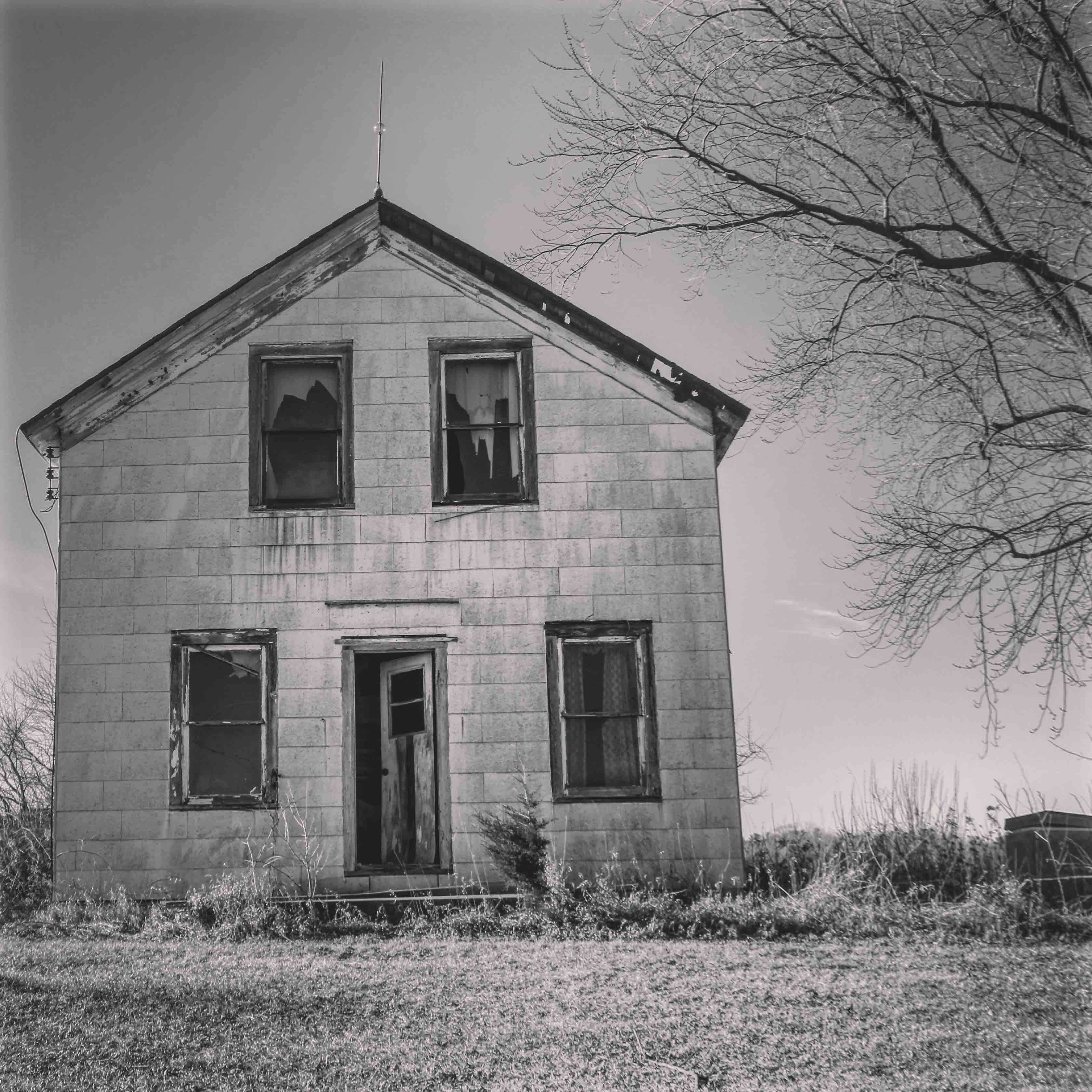 A weathered wooden house with a gabled roof and a chimney, surrounded by overgrown grass and a leafless tree.