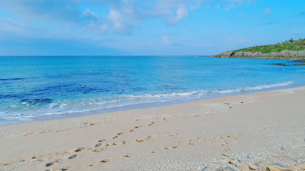 A beach with footprints in the sand, a clear blue sea, and a rocky outcrop with vegetation in the background.