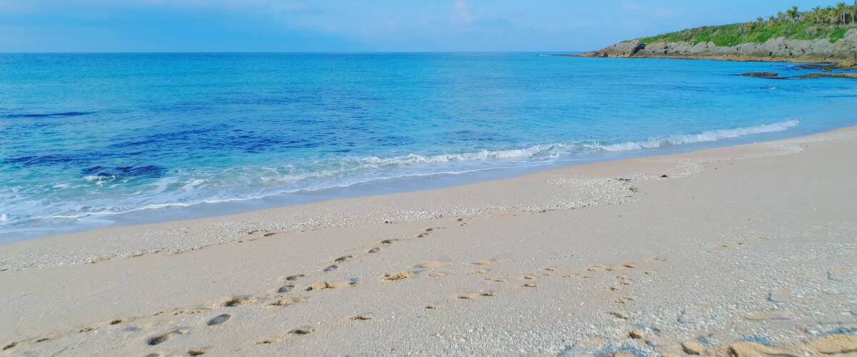 A beach with footprints in the sand, a clear blue sea, and a rocky outcrop with vegetation in the background.