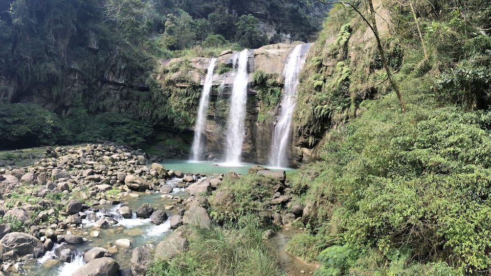 A waterfall cascading over rocks into a clear pool surrounded by lush greenery.